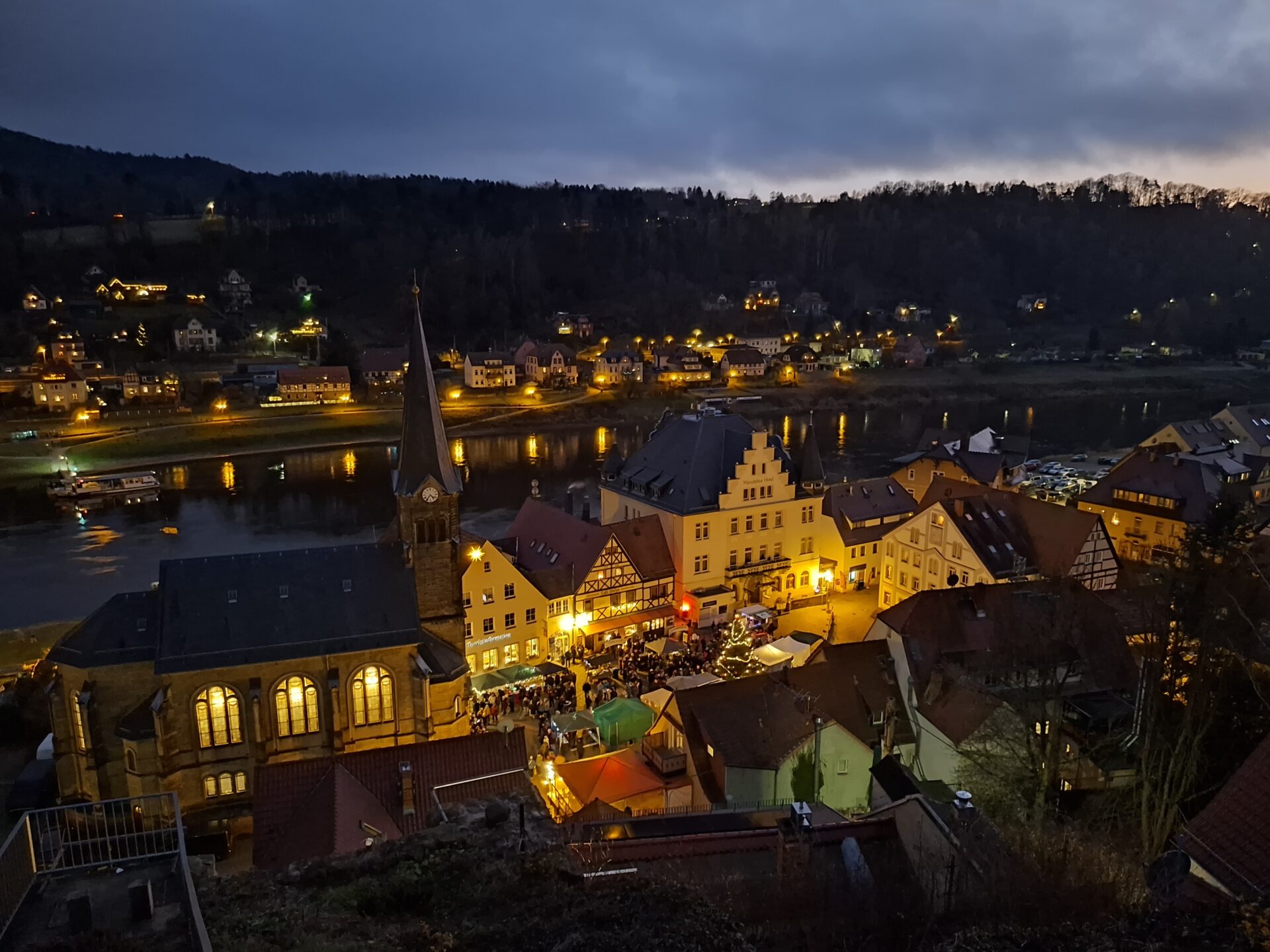 Blick auf den historischen Marktplatz der Stadt Wehlen an der Elbe bei Nacht. Lichter erhellen Fachwerk‑ und Altbauten sowie eine Kirche mit hohem Turm, im Hintergrund die Elbe und bewaldete Hügel.