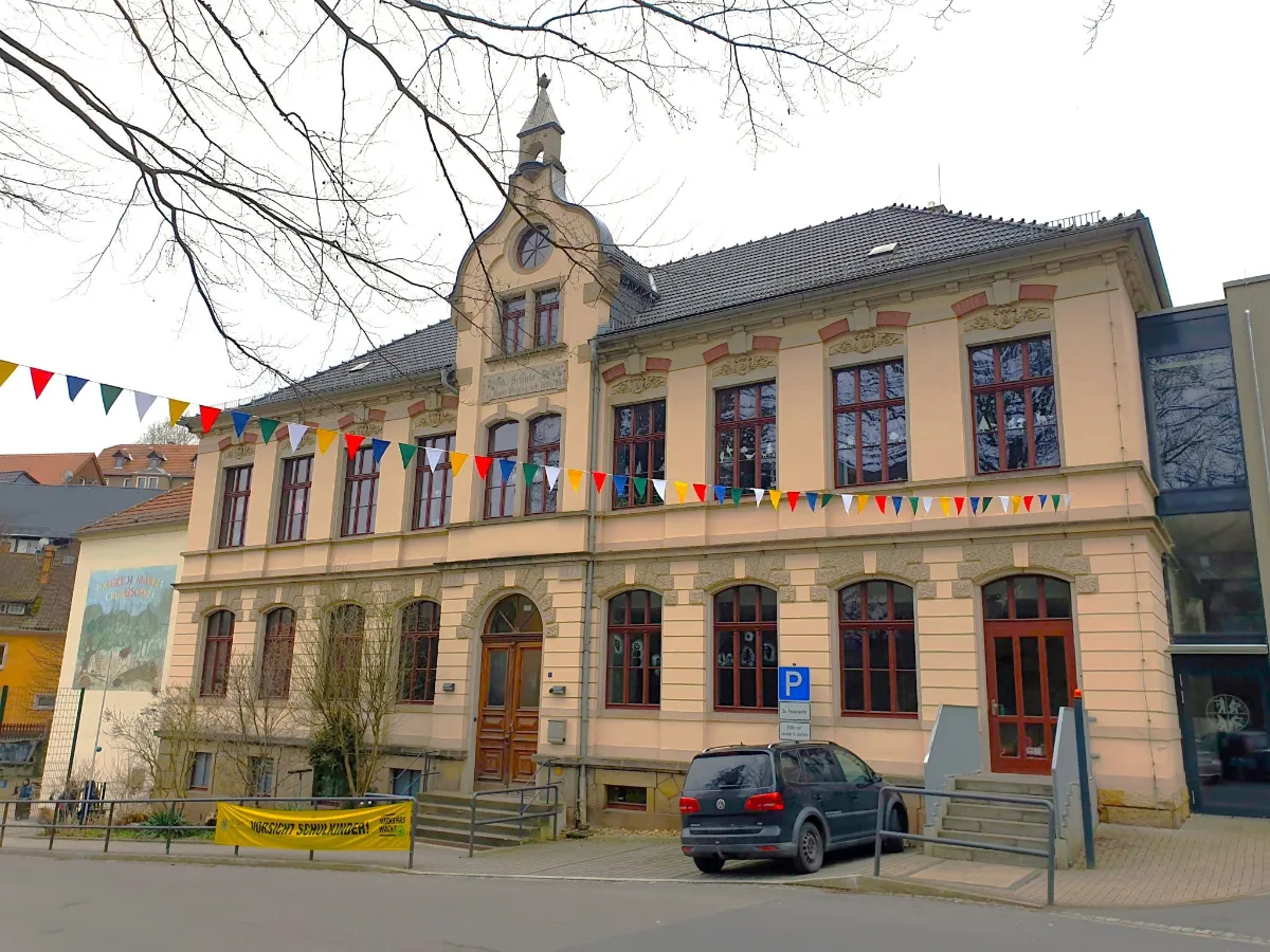Schulgebäude mit heller Fassade in Stadt Wehlen; über der Straße hängt ein Wimpel, an einem Geländer steht ein Banner mit der Aufschrift ‚Vorsicht Schulkinder‘, und auf dem Parkplatz neben dem Eingang steht ein Auto.