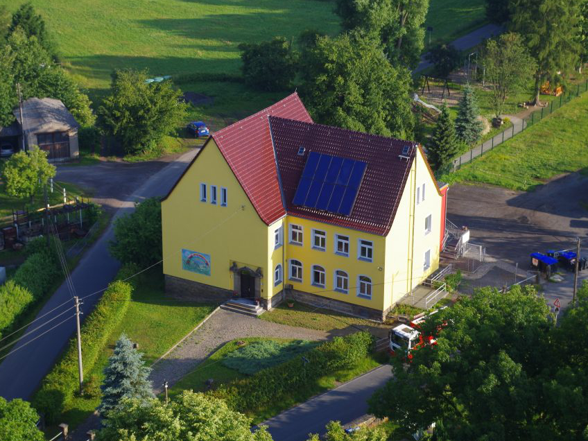 Luftbild eines Gebäudes mit gelbem Putz und rotem Dach, das die Kita Dorf Wehlen zeigt; vor dem Eingang sind ein Gehweg und bepflanzte Bereiche, im Hintergrund ein großer Spielplatz im Grünen.