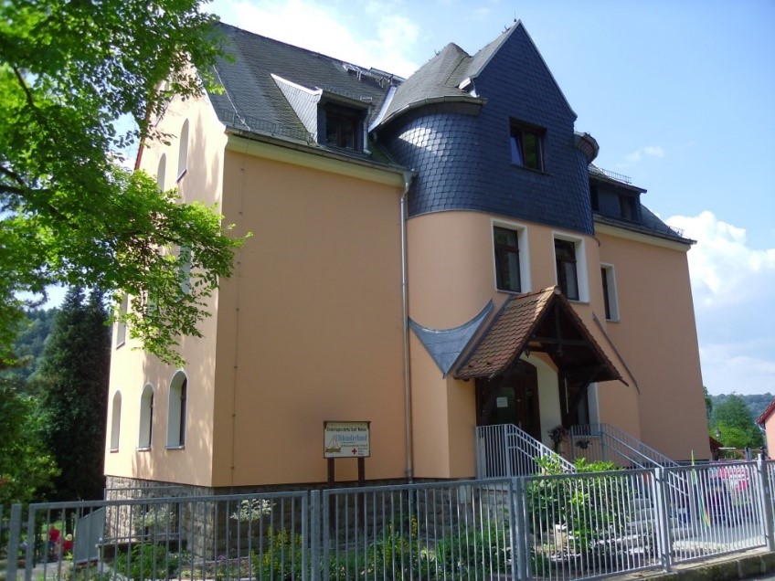 Ein Haus mit beigem Anstrich, das den Eingang der Kita Stadt Wehlen zeigt; neben der Eingangstreppe steht ein Schild, davor verläuft ein Zaun.