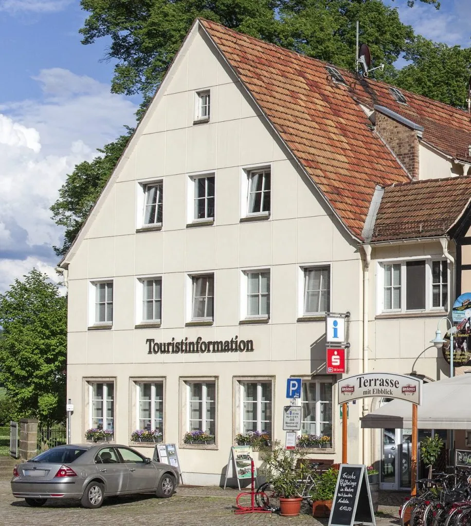 Helles Gebäude in Giebelansicht auf dem Markt in Stadt Wehlen mit vielen weißen Fenstern; an der Fassade steht "Touristinformation", davor steht ein Fahrzeug.