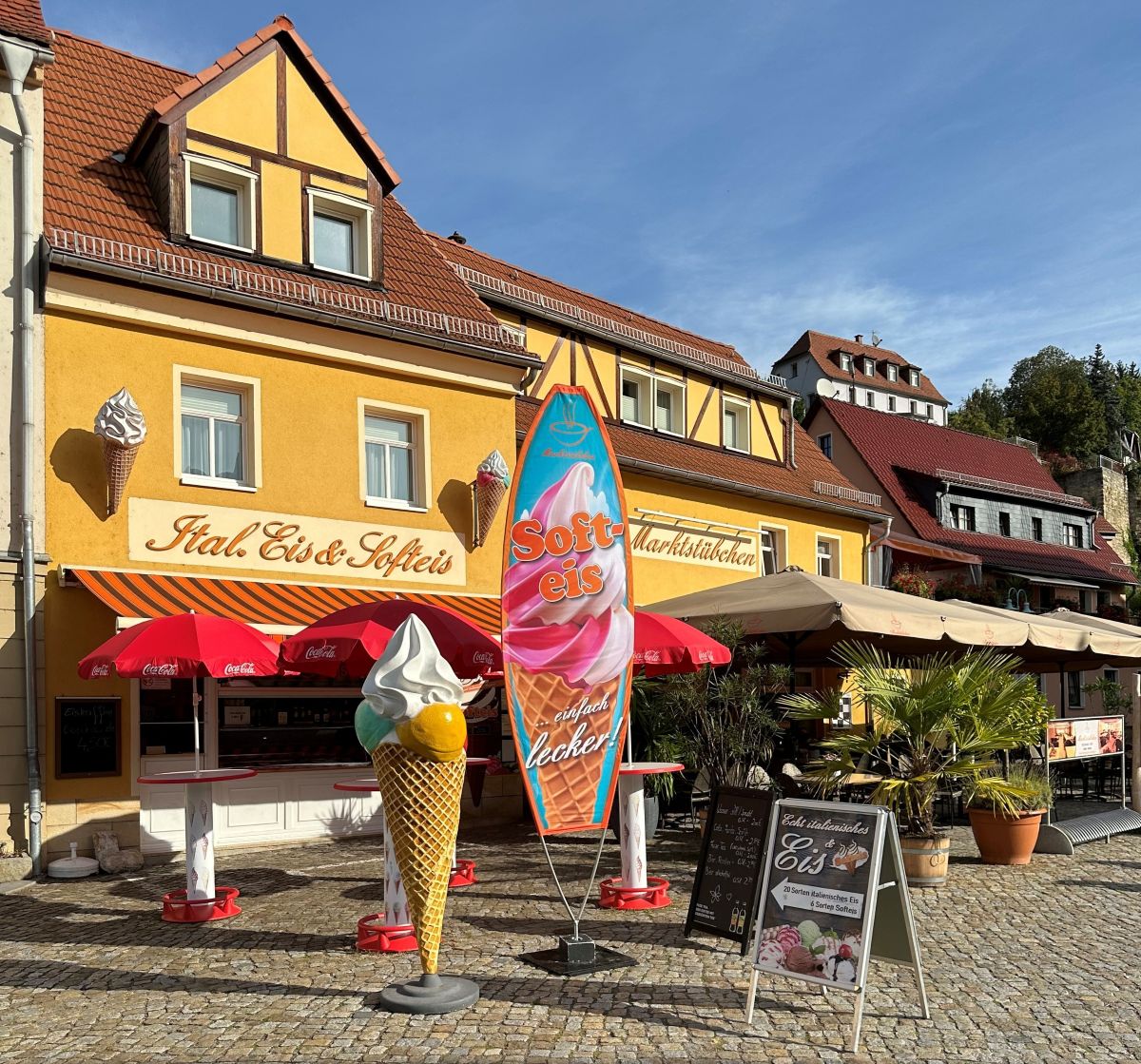 Nahaufnahme eines Tisches des Marktstübchens in Stadt Wehlen mit einem Espresso, einem Café Latte, Speisekarten und Pflanzen‑Dekoration; im Hintergrund unscharf der Eingang des Cafés und weitere Sitzplätze.